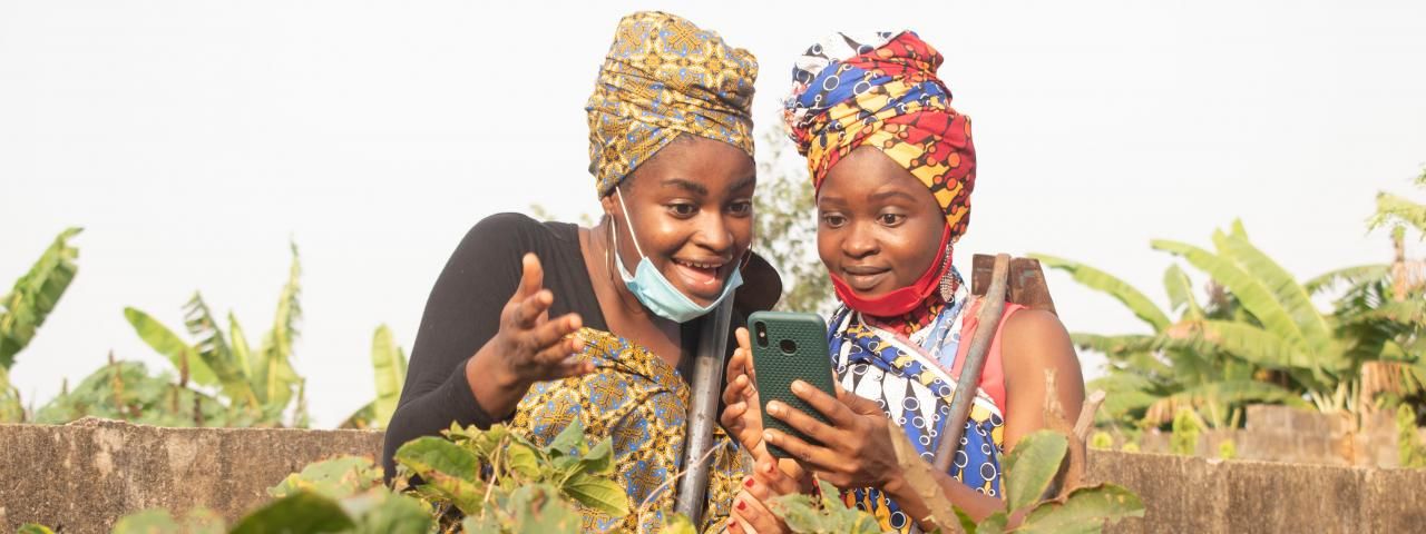 two ladies wearing african attire at their farmland, two ladies looking into their phone.